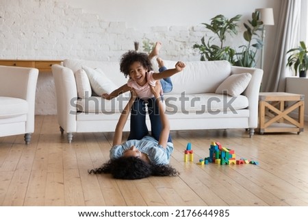 Carefree African family having fun, playing at home, mother lying on warm wooden floor lifts cute little daughter, girl spread her arms imagines herself flying in air indoor. Travel, leisure concept Royalty-Free Stock Photo #2176644985
