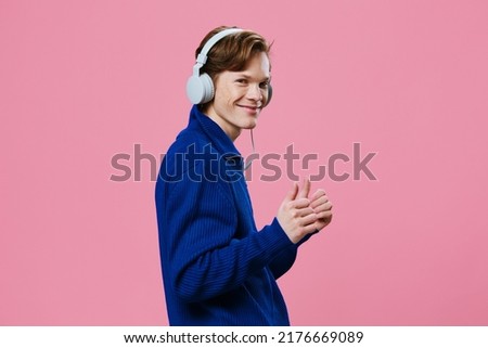 a joyful, happy man in a blue zip-up sweater dances to the music in his large, white, wired headphones on a pink background with space for text.