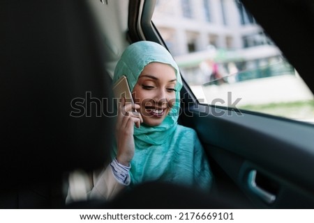 Beautiful Muslim Arabian woman with hijab sitting on backseat in luxury car. She using her smart phone and looking through window.