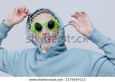 a close studio photograph of a man in a light blue hoodie, a mesh stretched over his face, standing on a light background, with bright green glasses holding a hoodie cord crossing his face with it