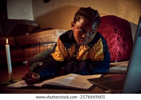 A young African school girl is doing homework on her laptop by candle light during load shedding power outage