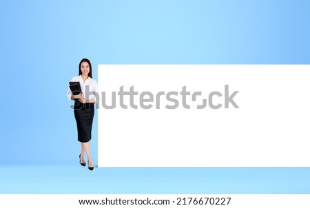 Attractive student wearing formal wear is holding notebook standing near blue wall with large empty mock up in background. Concept of imagination and inspiration for education, studying and learning