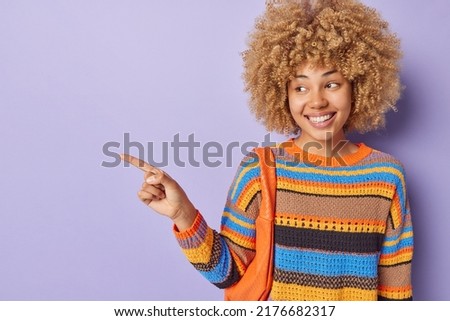 Positive good looking young woman points finger left shows place for your advertisement wears casual knitted sweater poses with bag against purple background. Look at this appealing product.