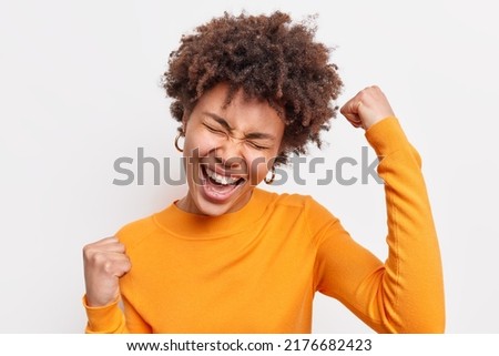 Joyful curly haired young woman makes yes gesture celebrates success clenches fists exclaims happily wears casual orange jumper feels very happy isolated over white background. Finally I did it Royalty-Free Stock Photo #2176682423