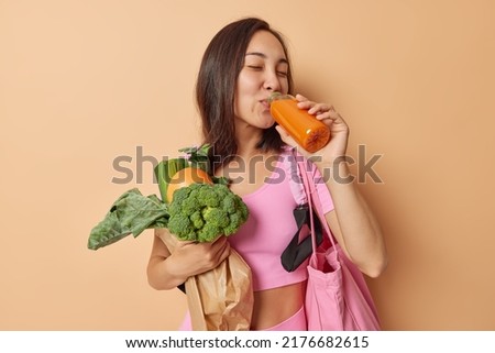 Diet and superfood concept. Healthy dark haired Asian woman drinks homemade orange juice detox drink carries fresh vegetables in paper bag has proper nutrition isolated over brown background Royalty-Free Stock Photo #2176682615