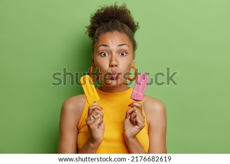 Temptation and chilling concept. Surprised curly haired youngwoman holds two appetizing ice creams keeps lips folded wears earrings and t shirt isolated over green background enjoys summer time Royalty-Free Stock Photo #2176682619