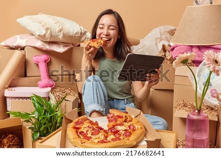 Cheerful Asian woman enjoys eating pizza and watching film on portable tablet poses on floor around cardboard boxes rents new apartment. Relocation at house. Tenancy and real estate concept. Royalty-Free Stock Photo #2176682641