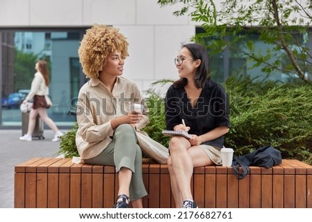 Happy Asian female journalist meets with famous person to take interview writes down notes in notebook drink takeaway coffee have pleasant conversation pose together against urban background Royalty-Free Stock Photo #2176682761