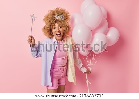 Festive occasion concept. Surprised glad woman with curly hair dressed casually prepares for party congraulates friend with anniversary holds magic wand ready to fullfil your dreams bunch of balloons