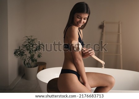 Woman taking care of body and skin, doing dry peeling massage with brush dressed in bikini, having beauty and spa procedure in luxury bathroom with modern bathtub on background