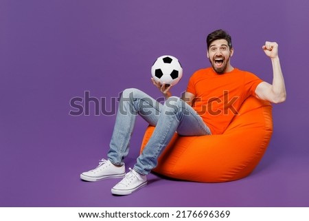 Full body young fan man he wear orange t-shirt cheer up support football sport team hold soccer ball watch tv live stream sit in bag chair do winner gesture isolated on plain dark purple background Royalty-Free Stock Photo #2176696369