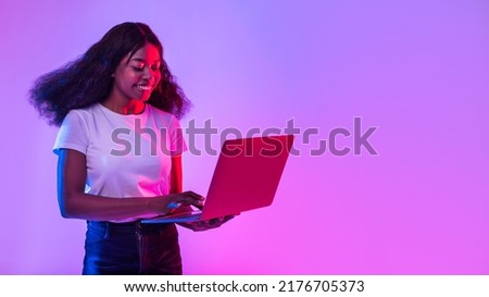Portrait of cheery young black woman using laptop pc, studying or working remotely, having online conference in neon light, banner with empty space. African American lady with computer browsing web Royalty-Free Stock Photo #2176705373