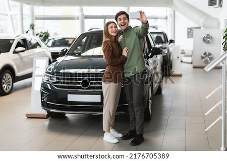Full length of cheery young couple taking selfie on mobile phone near their new car at auto dealership, copy space. Joyful millennial spouses photographing themselves at showroom, buying automobile