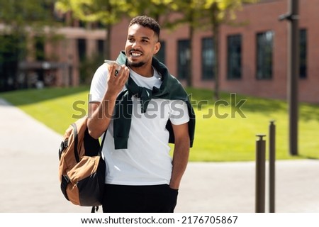 Positive stylish black guy talking on smartphone, using loudspeaker or recording audio message, walking down in urban city area, copy space. African american man speaking outdoors