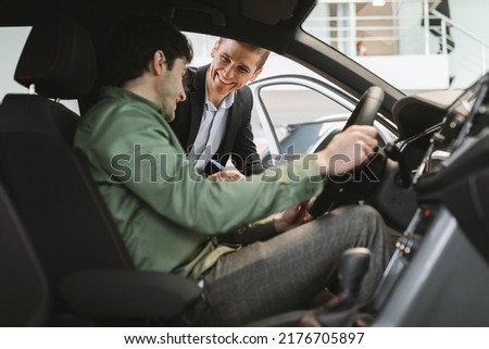 Happy millennial man buying new vehicle, clarifying car purchase details with salesman, sitting inside auto salon at dealership center. Young Caucasian guy purchasing automobile at showroom Royalty-Free Stock Photo #2176705897