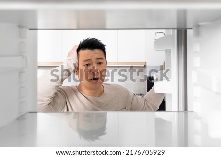 Shocked middle aged chinese man looking inside empty fridge and touching his head, lack of food, need for shopping, suffering from financial hangover while pandemic, shot from inside, copy space Royalty-Free Stock Photo #2176705929
