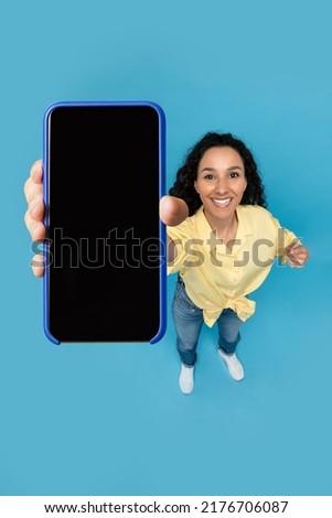 Pretty young lady pointing at smartphone with blank screen on blue studio background, mockup. Millennial woman recommending new mobile app, advertising website, copy space. Above view