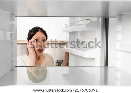 Shocked young asian woman looking inside empty fridge and touching her face, lack of food, need for shopping, suffering from financial hangover while pandemic, shot from inside, copy space Royalty-Free Stock Photo #2176706091