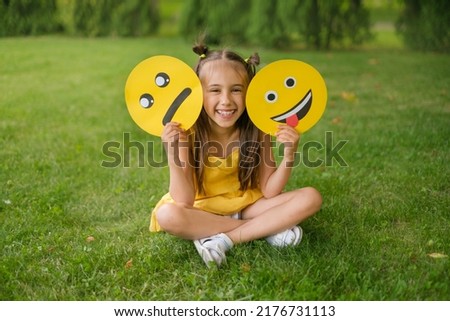 A laughing, cheerful child holds two emoticons in his hands - a sad, upset one and a smile face showing his tongue
