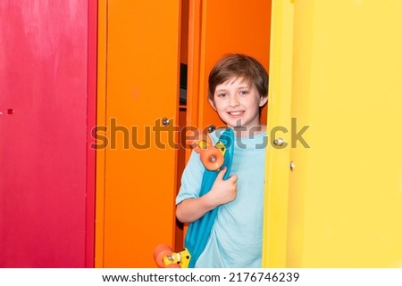 a smiling boy holds a skateboard, stands near bright multi-colored school lockers