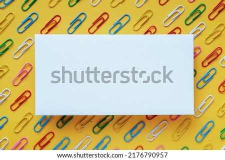 White blank paper card on background of multi-colored paper clips on yellow background. Template for design, space to insert text or logo. Minimalistic design. Top view