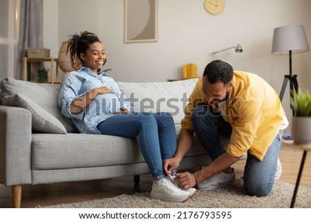 Pregnancy Lifestyle. Loving African American Husband Helping Pregnant Wife Lacing Her Shoes While She's Sitting On Sofa Touching Belly At Home. Support And Care, Family Concept