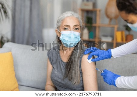Doctor in protective gloves makes injection of european old woman in mask with gray hair in living room interior. Vaccination for health care, immunization against flu and covid-19 virus due outbreak