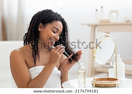 Happy Black Lady Applying Moisturising Oil On Damaged Hair Ends After Shower, Smiling African American Woman Sitting Near Mirror In Bathroom Interior, Making Haircare Routine At Home, Closeup Royalty-Free Stock Photo #2176794181