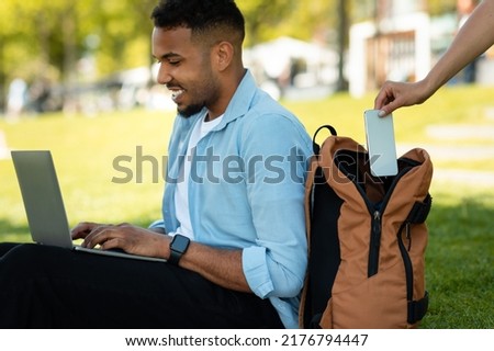 Pickpocket thief stealing cellphone from man's backpack while african american guy working online on laptop computer, sitting on grass in park Royalty-Free Stock Photo #2176794447