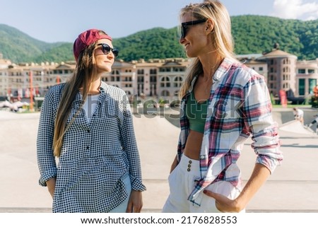 Two young cheerful girlfriends are talking and laughing on a summer morning in a skatepark