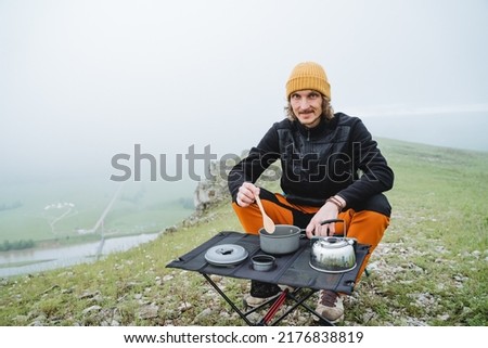 A tourist guy sits at a table in the mountains having breakfast, a man joyfully dining in nature on a mountain hike, cooking in the forest, foggy weather is cold. High quality photo