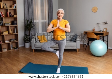 Yoga practice on retirement. Calm senior lady standing in tree pose, keeping balanced, exercising on yoga mat at home. Happy aged woman training indoors, copy space