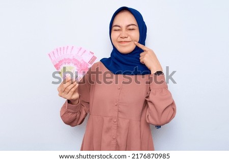 Excited young Asian muslim woman in pink shirt holding money banknotes isolated over white background. People religious lifestyle concept