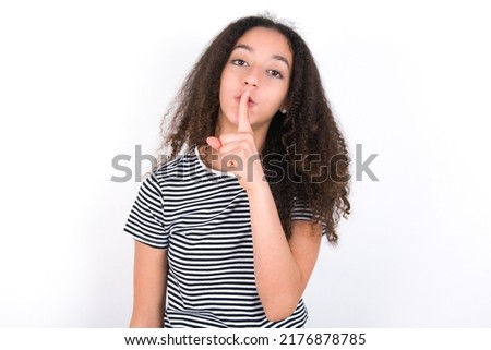 Surprised young beautiful brunette woman wearing striped t-shirt over white wall makes silence gesture, keeps finger over lips and looks mysterious at camera