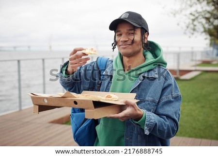 Happy black guy in casualwear holding slice of pizza over square box while having snack and looking at camera during picnic in park