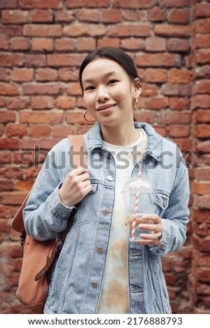 Young pretty woman in casualwear holding backpack on shoulder and soda in hand while standing against brick wall of building