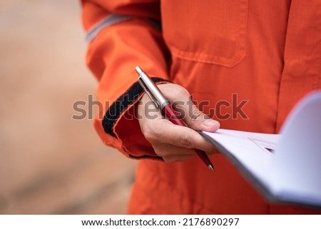 A supervisor in orange coverall is holding luxury ballpoint pen, action to review the paper document during perform safety audit. Industrial and business working scene photo. Selective focus.