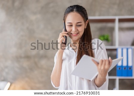 Young smiling Asian woman talking on mobile phone, standing modern office with a document in her hand