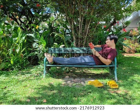 Young Asian man wearing glasses with black hair holding a book sitting with legs stretched on a bench. happy and relax. What are thinking or reading notebook in the summer park city