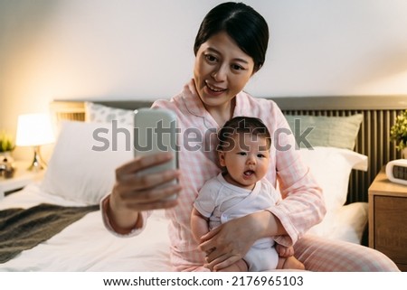 happy asian mother wearing pajamas holding and looking at smartphone screen is taking a selfie of herself and her lovely baby in arms in the bedroom at night.