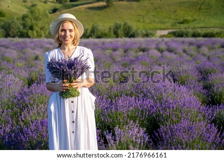 woman model in white dress outfit with hat is standing in lavender field, photo session. Young girl is holding lavender bouquet. Portrait 