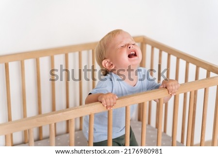 Portrait of upset sad frustrated one year old baby boy getting hysterical standing in bed asking to pick him up, seeking attention of parents crying out loud. Child temper tantrum Royalty-Free Stock Photo #2176980981
