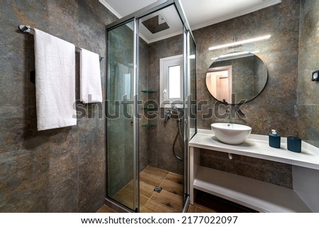 Modern bathroom in grey and brown colors with glass shower cabin in the corner, tap mounted in the wall, round mirror, white bowl sink. Selective focus.