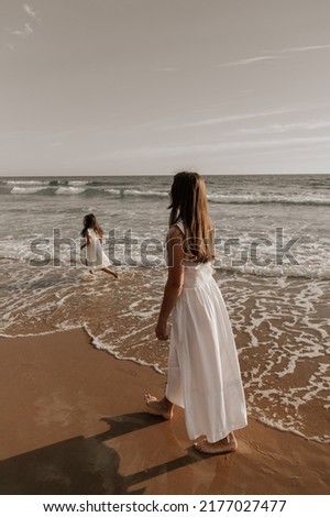 Full body barefoot mom in white dress walking on wet sand near child playing in foamy sea waves on cloudy day