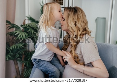 Pregnant swedish woman in beige t-shirt sitting with her daughter in light blue chair, little girl kissing her mom in a forehead. Mom and daughter with tails at home, Maternity and pregnancy concept.