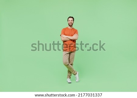 Full body young smiling fun man 20s wear casual orange t-shirt isolated on plain hold hands crossed folded look camera pastel light green color background studio portrait. People lifestyle concept