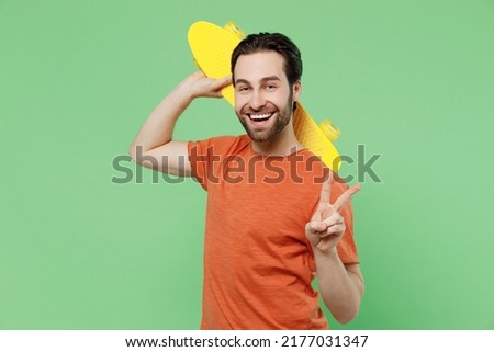 Young smilign happy cool man 20s in casual orange t-shirt hold yellow scateboard pennyboard show v-sign isolated on plain pastel light green color background studio portrait. People lifestyle concept
