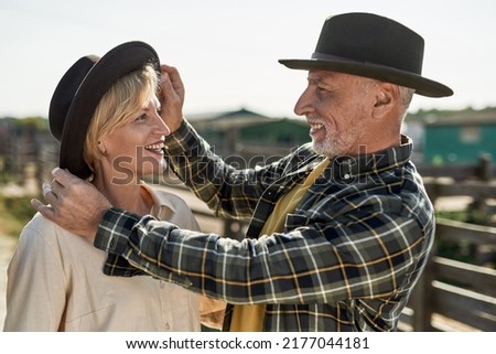 Grey hair male farmer correcting hat on head of his blonde adult wife on farm or ranch. Smiling caucasian man and woman spend time together. Modern countryside lifestyle. Agriculture. Sunny day Royalty-Free Stock Photo #2177044181