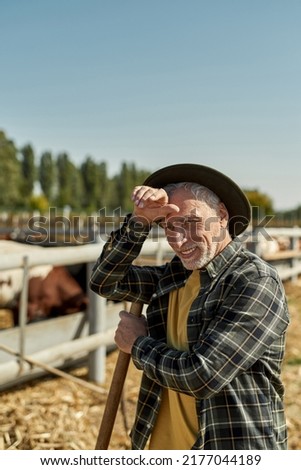 Tired elderly male farmer with agricultural tool resting and looking away during work on farm. Caucasian man wearing cowboy hat. Modern countryside lifestyle. Agriculture and farming. Sunny day