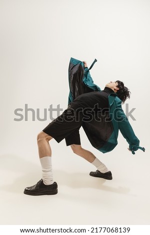 Portrait of stylish young man in black outfit and green coat posing isolated over grey studio background. Chaotic movements. Concept of modern fashion, art photography, style, queer, uniqueness, ad